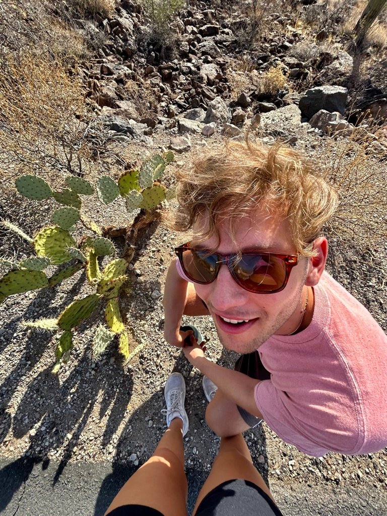 A cheerful young man wearing sunglasses looks up smiling next to a cactus on a desert trail, holding hands with someone whose feet are in frame.