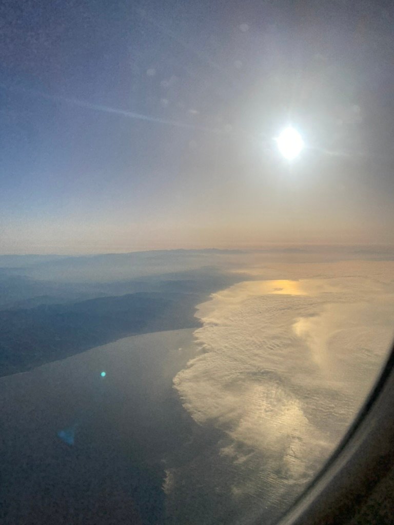A view from an airplane window showing the sun shining over coastal clouds and ocean, with hazy landforms fading into the distance.