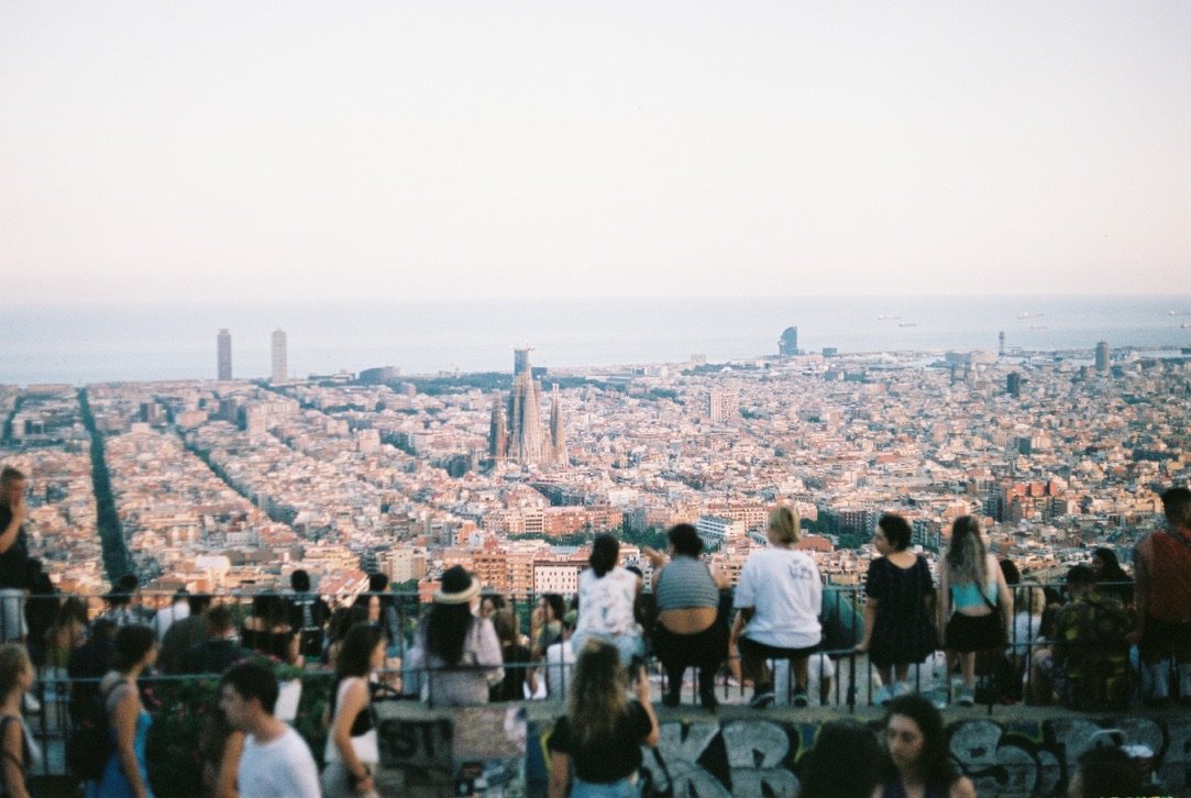 A group of people sit and stand at a scenic viewpoint overlooking the city of Barcelona, with the sea and skyline stretching into the distance at sunset.