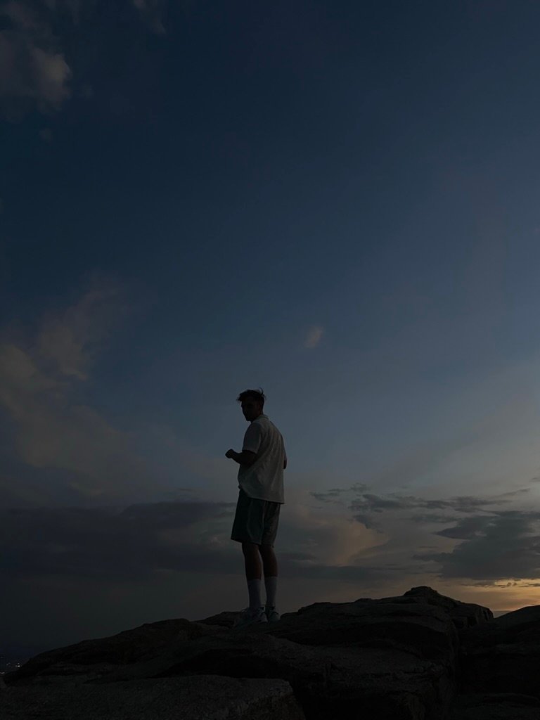 A person stands alone on a rocky mountaintop at dusk, silhouetted against a vast, peaceful sky filled with fading light and clouds.