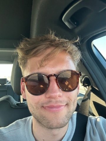 A young man wearing sunglasses smiles while sitting in the driver’s seat of a car, with sunlight shining in from the open road behind him.