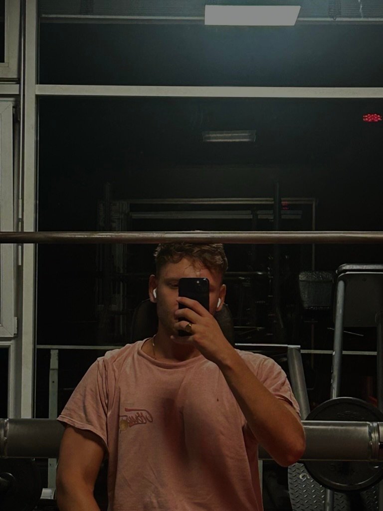 Man sitting on a bench press in the gym, holding a phone, wearing workout clothes and earbuds