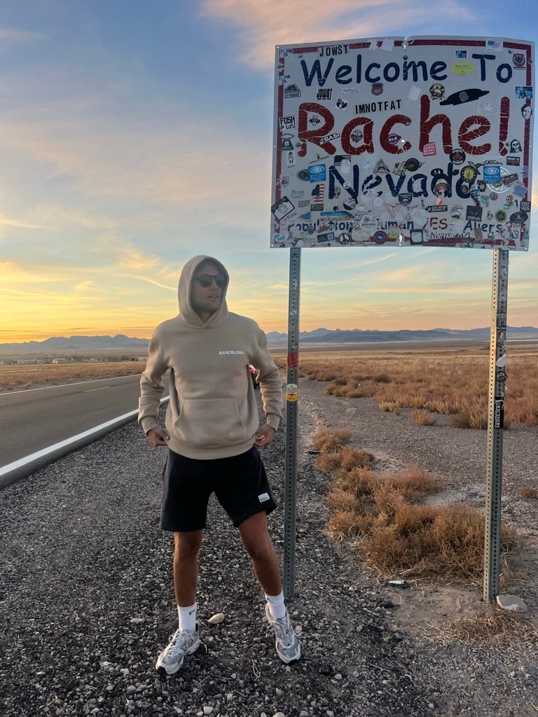 A man stands confidently under a sticker-covered "Welcome to Rachel, Nevada" sign along a quiet desert road at sunrise, wearing a hoodie and sunglasses.