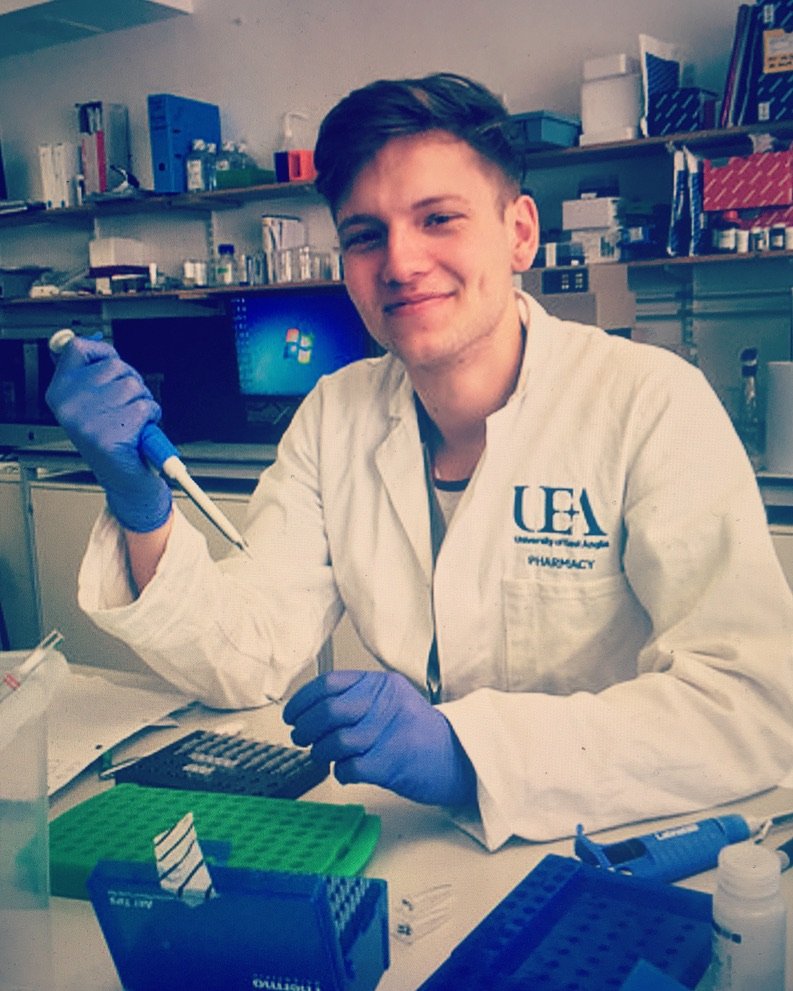 A young man in a white lab coat and gloves smiles while working at a laboratory bench, appearing focused and prepared.