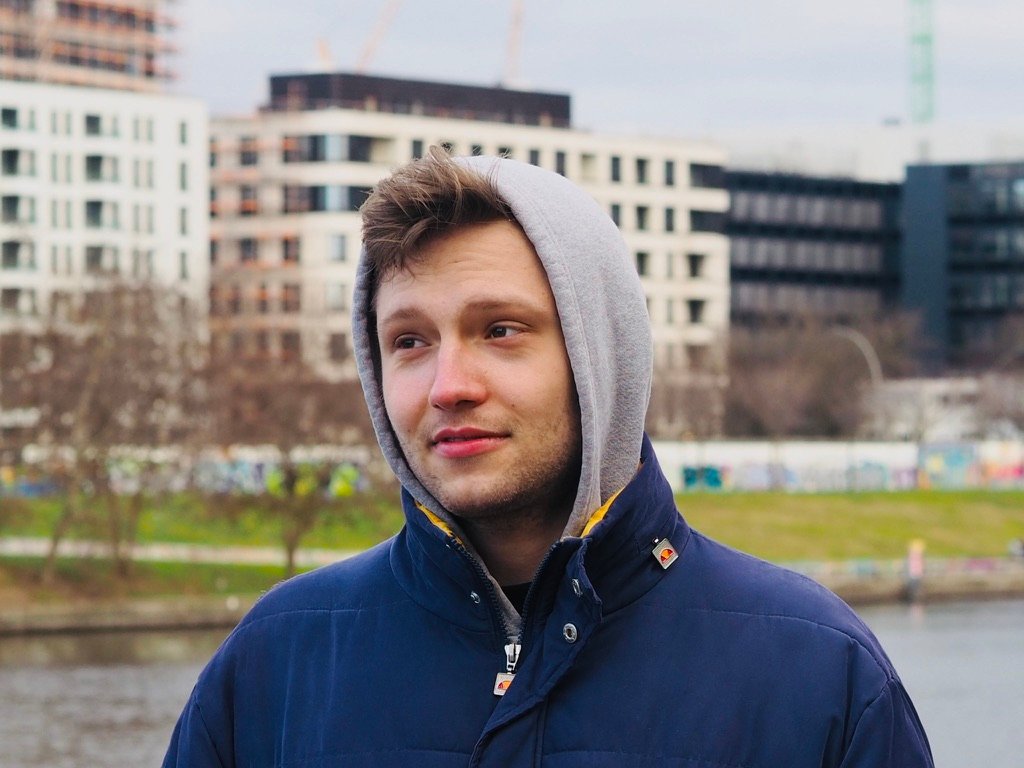 A young man in a hoodie and winter jacket looks off thoughtfully in an urban park setting, with modern buildings behind him and water nearby.