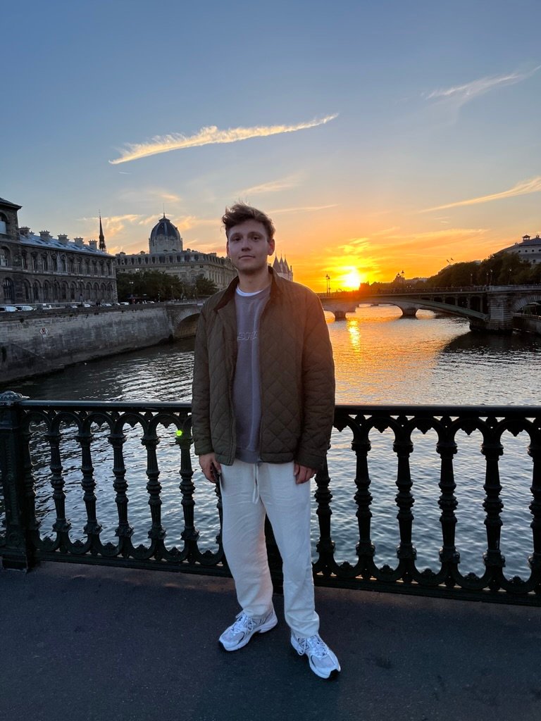 A young man stands calmly on a Paris bridge during sunset, with the river and historic buildings glowing in the golden light behind him.