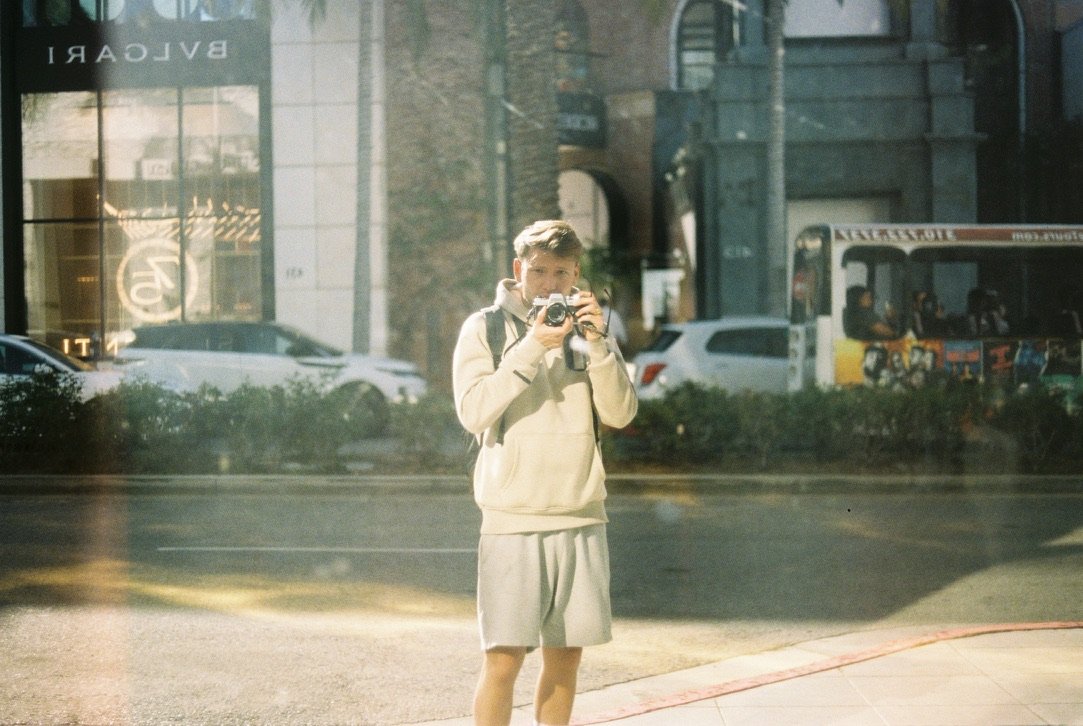 Man taking a photo with an analog camera, reflected in a shop window on a street in Hollywood, Los Angeles