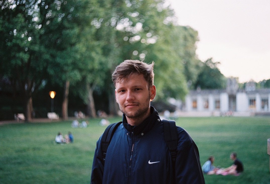 A young man stands calmly in a park with trees and grass behind him, wearing a navy jacket and backpack, with people relaxing in the background.