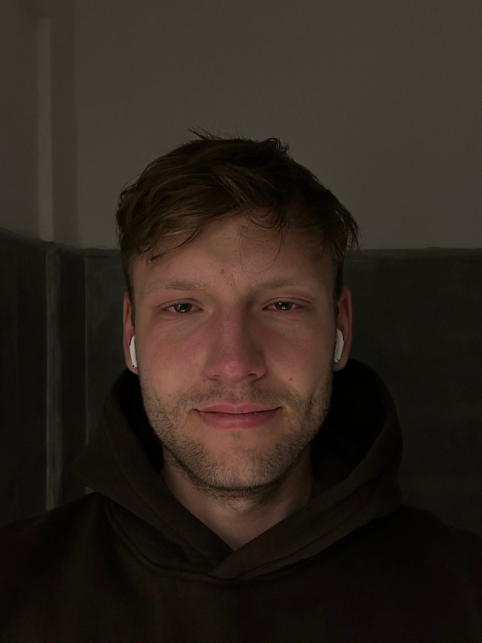 Man in hoodie with headphones, calm and slightly tired, standing in dim light