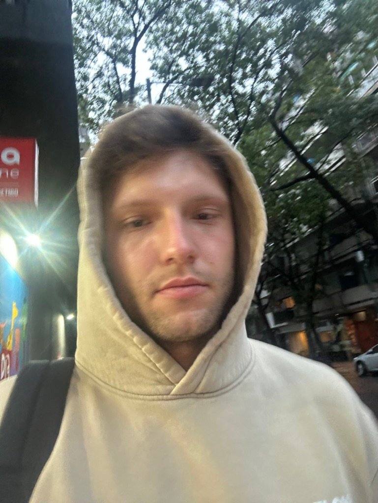 A young man in a tan hoodie walks through a city street at dusk, looking solemn with slightly blurred surroundings and dim lighting.