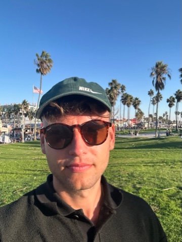 A young man in sunglasses and a cap stands on a grassy field with palm trees and blue sky in the background, looking calmly into the camera.