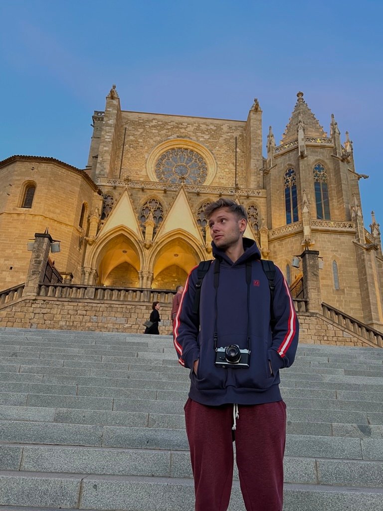 A young man stands on stone steps in front of a grand cathedral at sunset, wearing a hoodie and camera, looking upward thoughtfully.