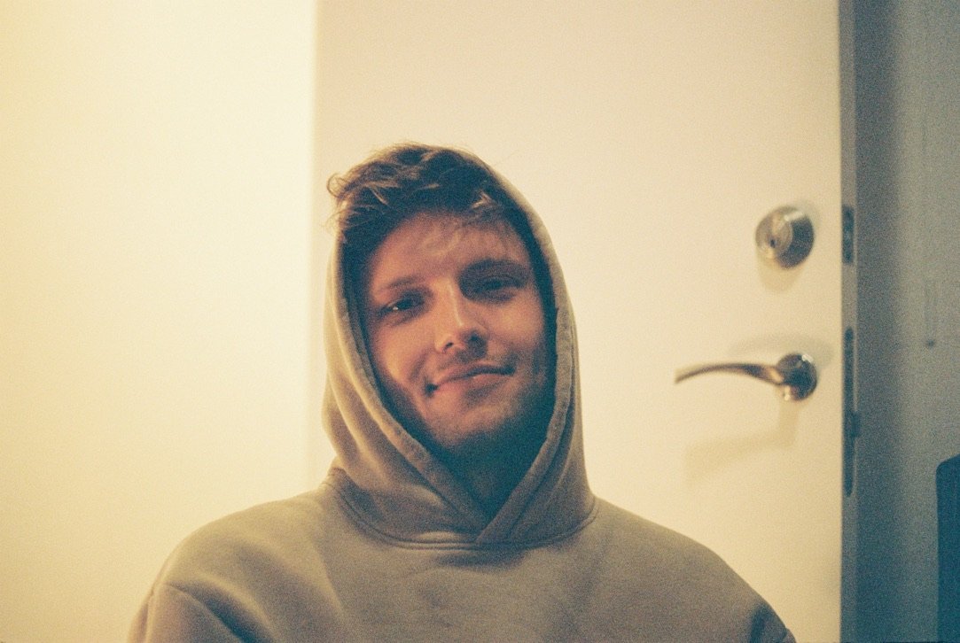 Young man in a hoodie sitting near a door, faint smile, peaceful but reflective expression