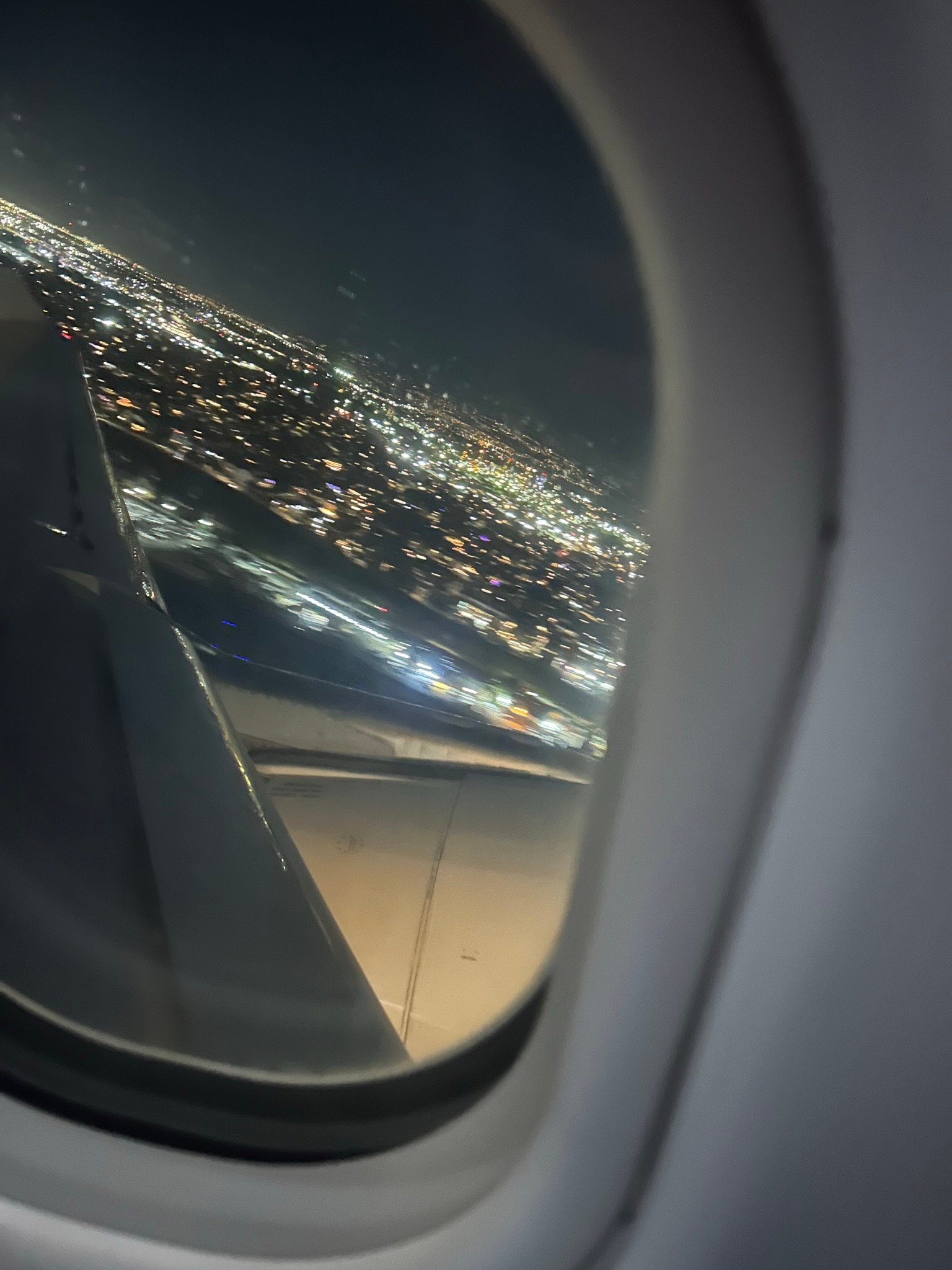 A nighttime view from an airplane window showing glowing city lights below and part of the plane wing illuminated in the dark sky.