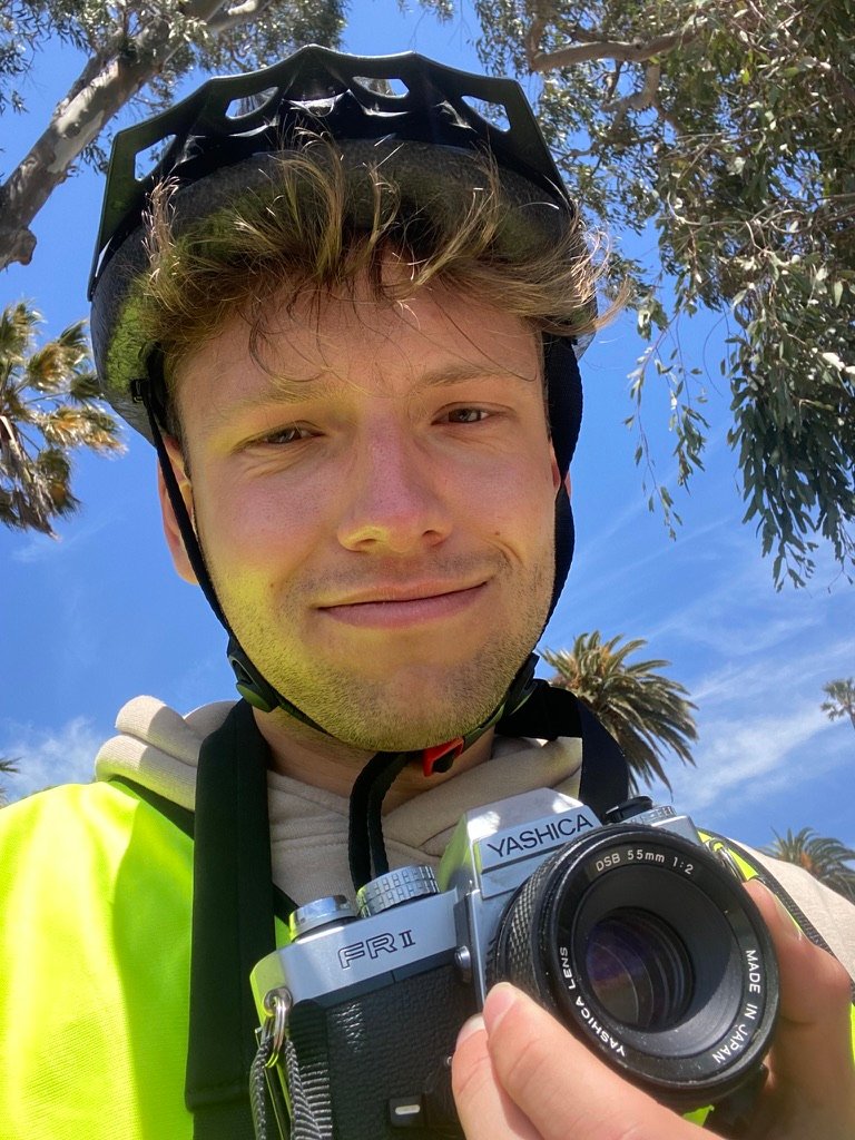 A smiling young man wearing a helmet and holding a vintage camera looks into the lens under a clear blue sky, with trees and palm leaves in the background.