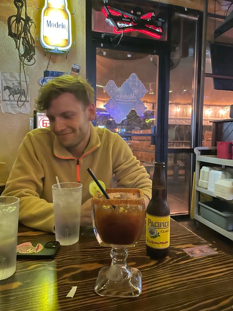A young man sits at a restaurant table with drinks and a slight smile, under warm lighting and a casual atmosphere.