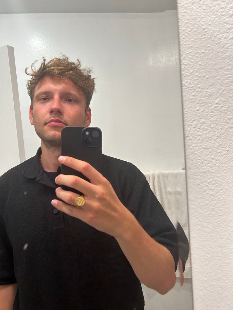 A young man takes a mirror selfie in a white bathroom, wearing a black shirt and gold ring, looking calmly into the lens.