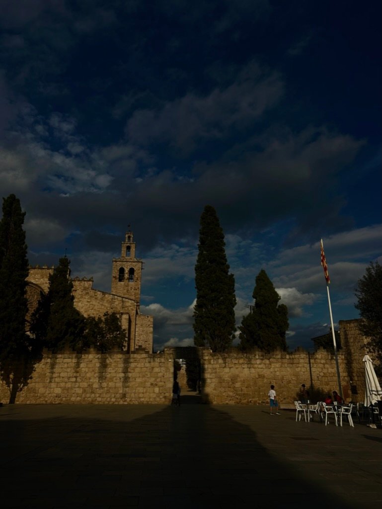 Stone courtyard with a medieval church tower and tall cypress trees under a dramatic evening sky.