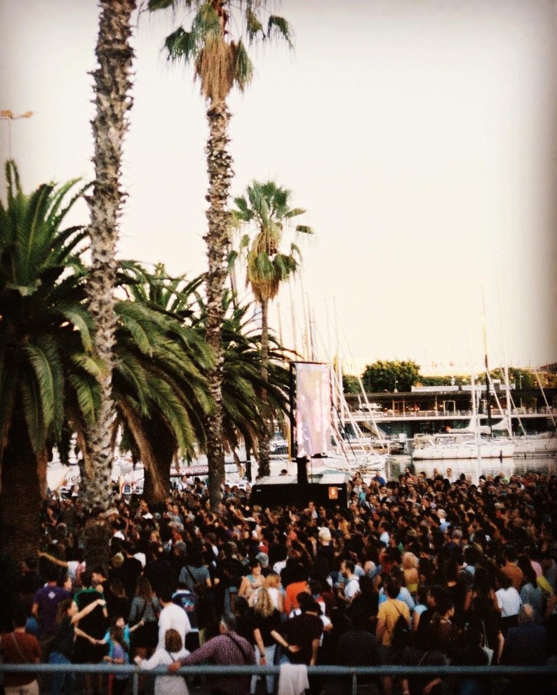 A large crowd gathers under tall palm trees near a marina at sunset, with a screen visible among the people.