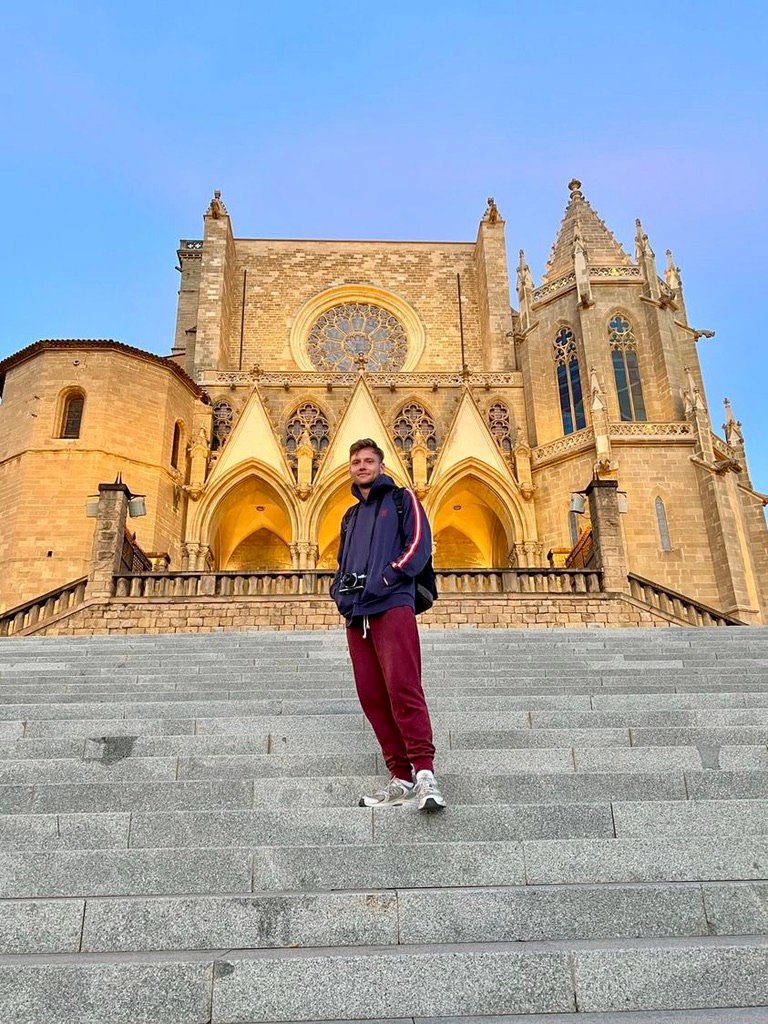 Man standing on the stone steps of a grand cathedral, facing the camera as the sky glows in early morning or evening light.