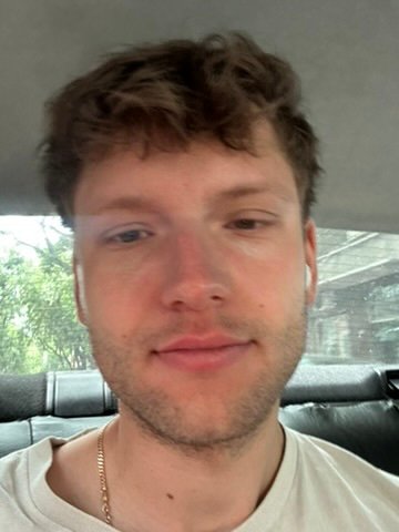 A young man with tousled brown hair sits in the backseat of a car, wearing a white shirt and AirPods, with a soft expression on his face