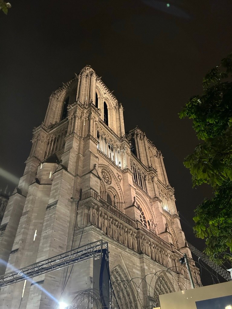 Nighttime view of the illuminated Notre Dame Cathedral in Paris, framed by trees and scaffolding.