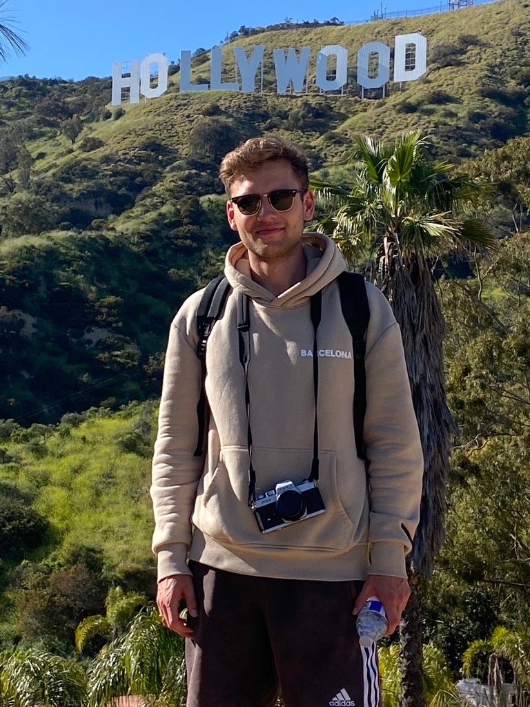 A young man in sunglasses and a hoodie stands smiling in front of the Hollywood sign, with trees and hills behind him and a camera around his neck.
