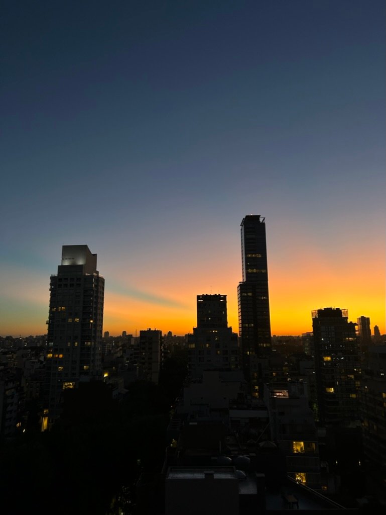 A city skyline at twilight with tall buildings silhouetted against a radiant orange and blue sunset