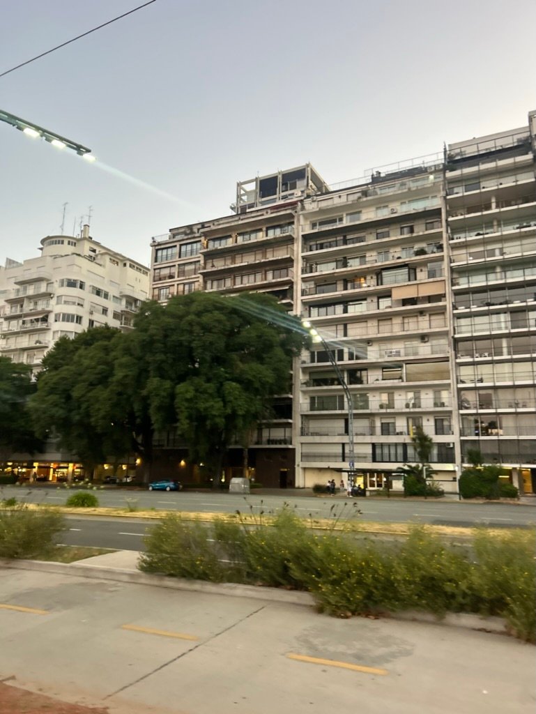 A street view of a mid-rise residential building in soft evening light, with trees and streetlights nearby