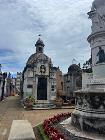 A historic cemetery with ornate mausoleums, stone statues, and flower-lined paths under a cloudy sky
