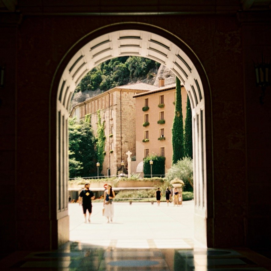 View through a large stone archway framing a courtyard with tall trees and a historic building in warm sunlight.
