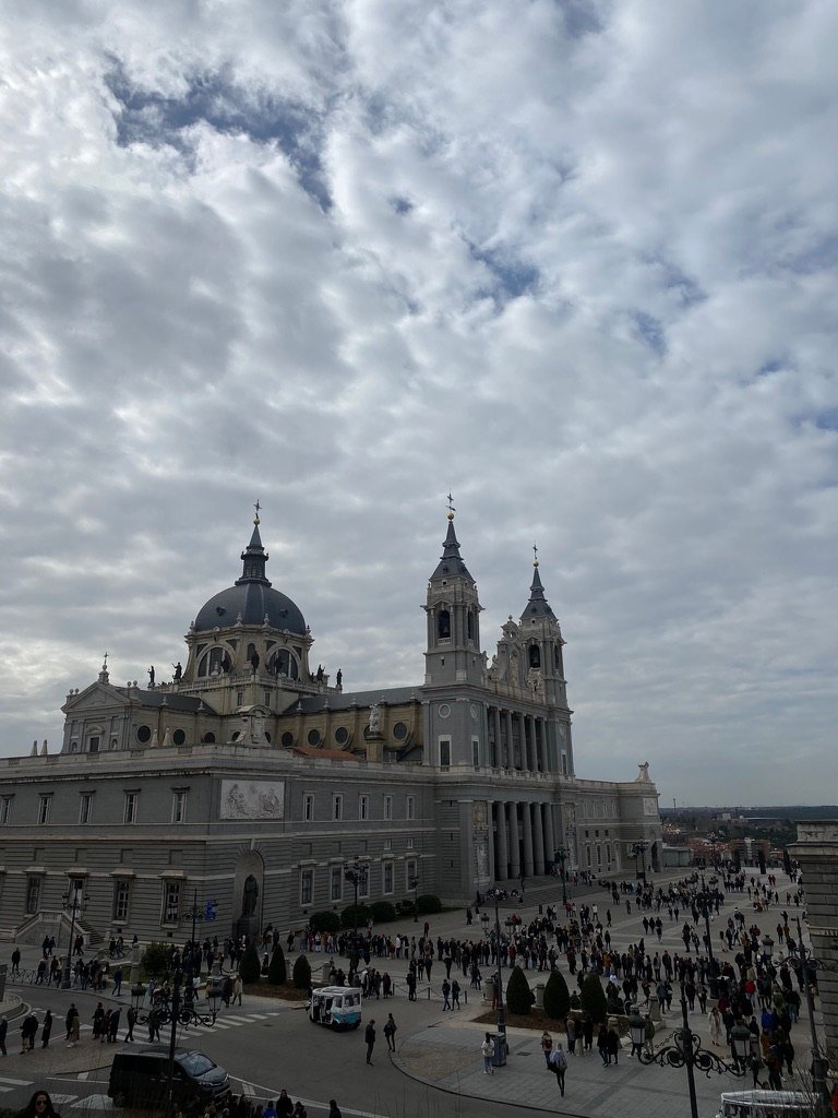 A grand cathedral with domes and towers stands under a dramatic, cloud-filled sky, with crowds of people gathered in the large courtyard below.