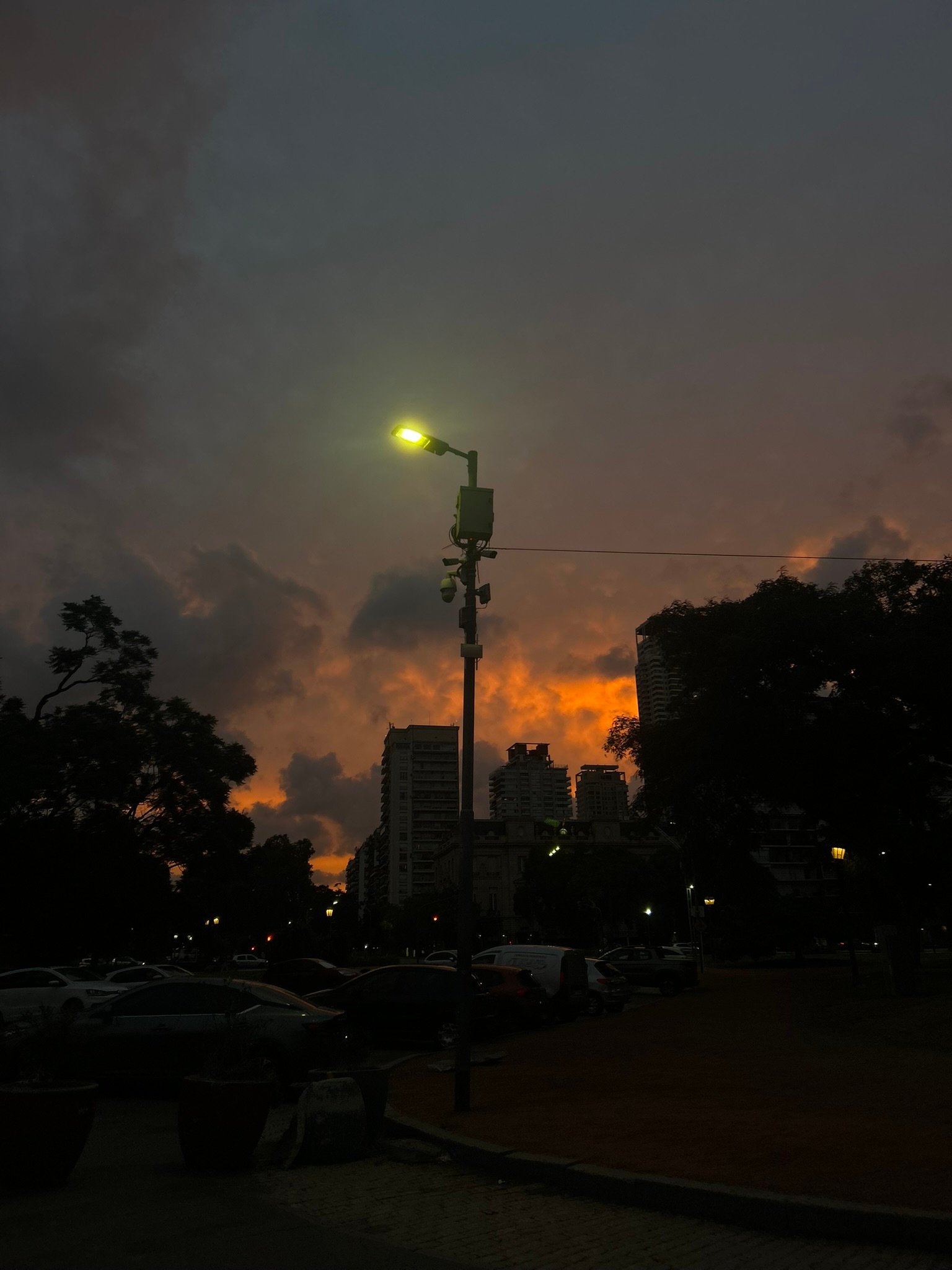 A city street at dusk with a single streetlamp glowing greenish-yellow against dramatic orange clouds in the sky
