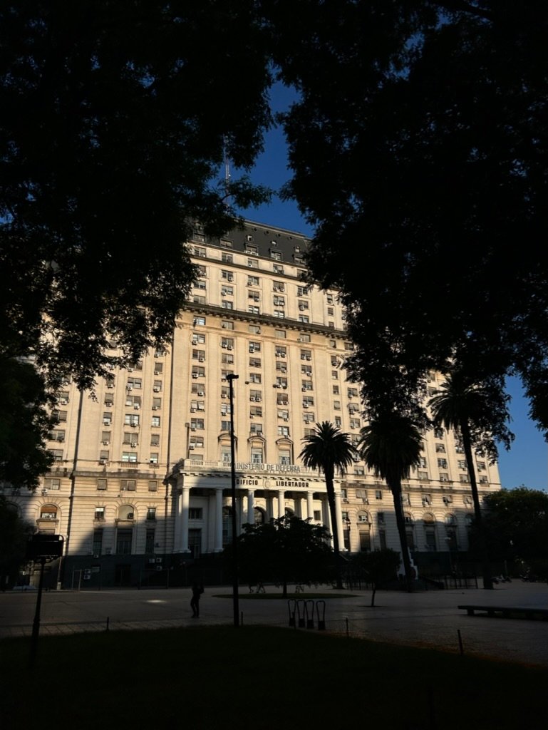 A tall government building partially illuminated by sunlight, framed by tree shadows in the foreground.