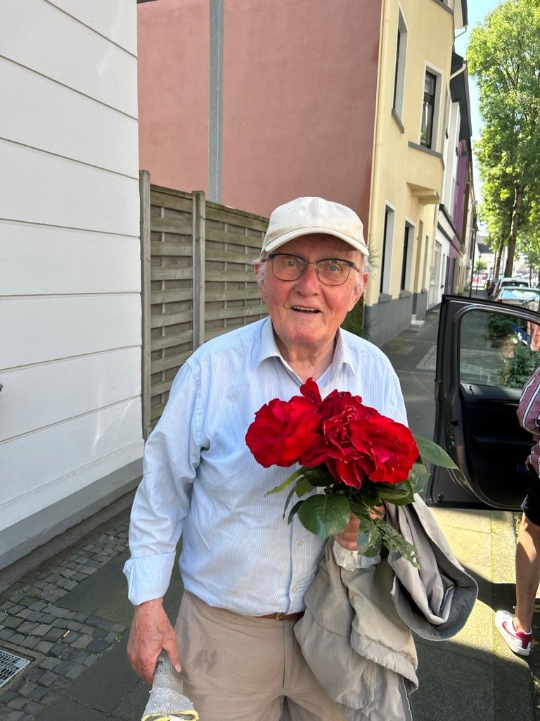 An elderly man in a white cap holding a bouquet of red roses, smiling on a sunny sidewalk near pastel-colored houses.