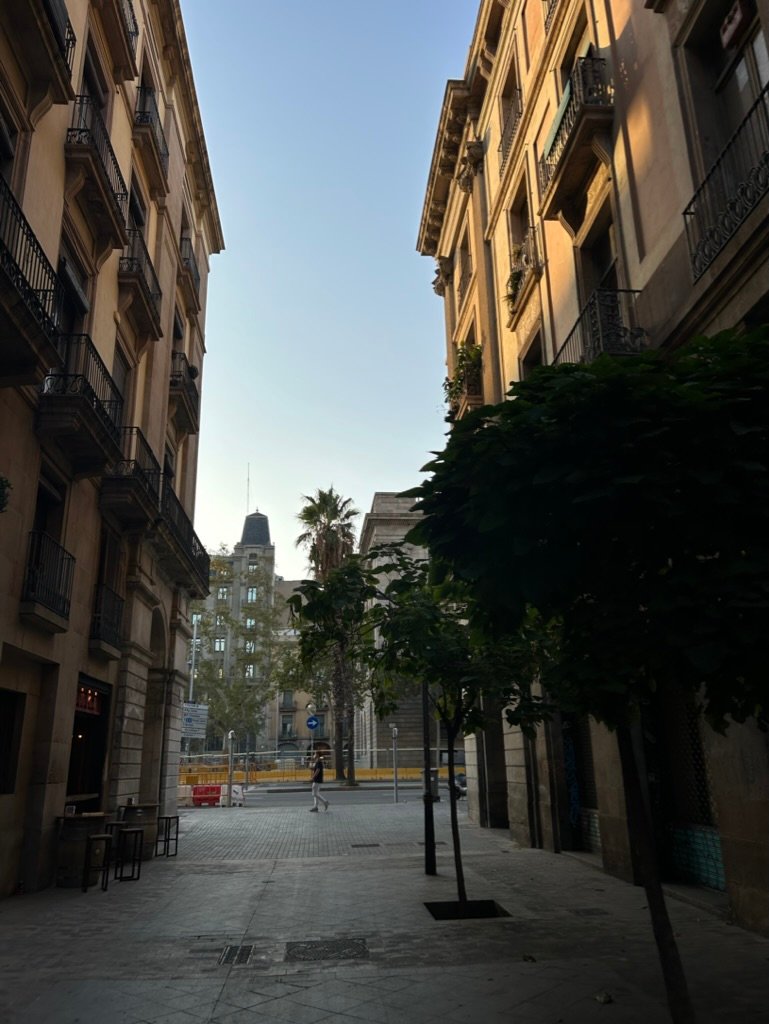 A narrow European street at sunrise, flanked by tall, historic buildings with iron balconies and soft morning light.