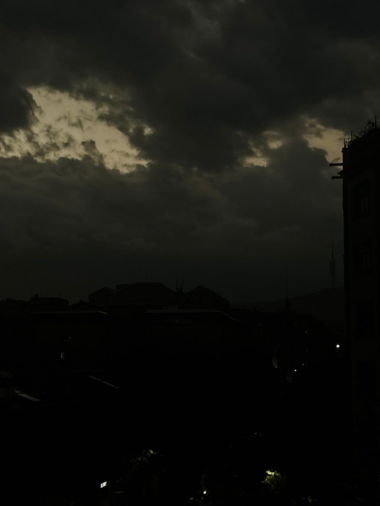 A dark, moody skyline with heavy clouds and faint city lights beneath a break in the clouds revealing dim light.