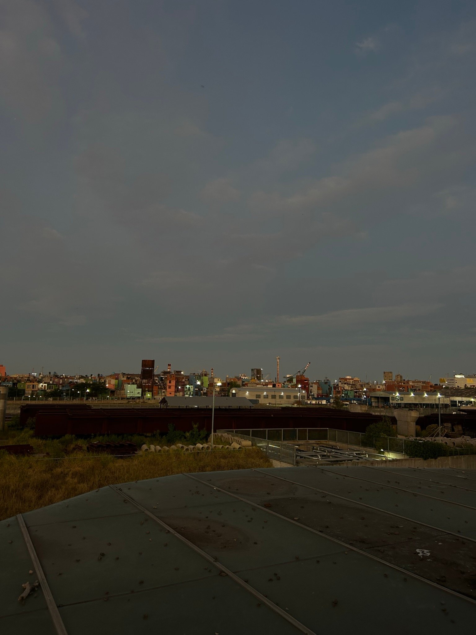A dimly lit industrial area at dusk with a cloudy sky, scattered buildings in the distance, and faint city lights beginning to glow