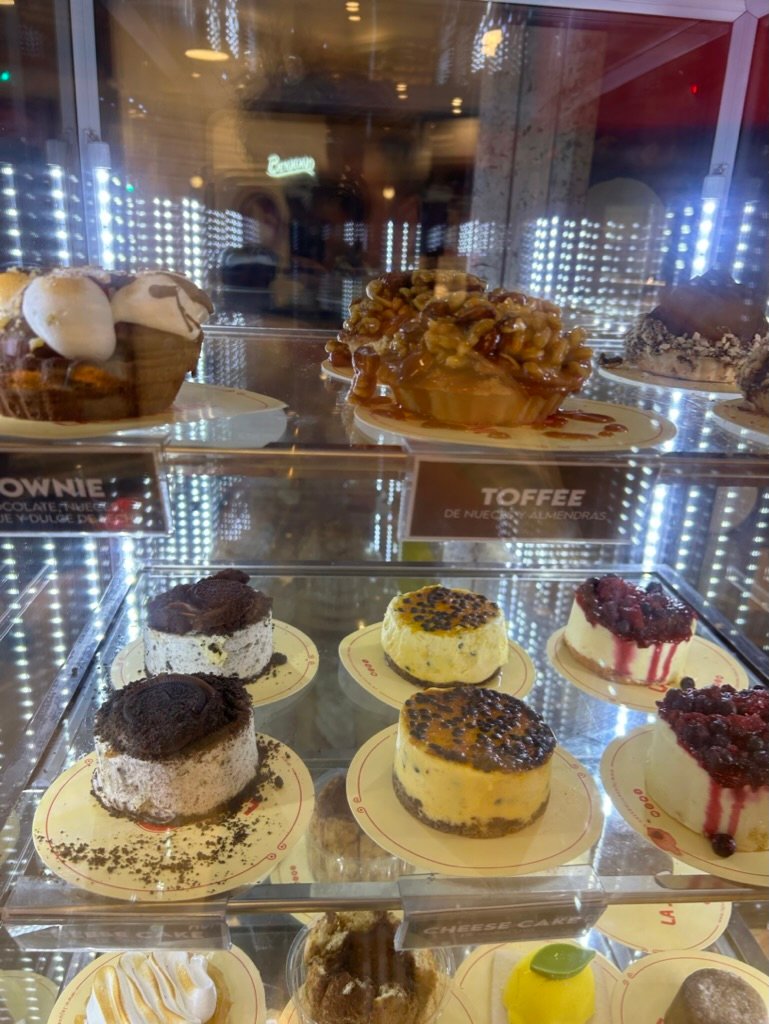 A brightly lit dessert case filled with various cheesecakes, toffee tarts, and brownie treats, seen through glass with reflections.