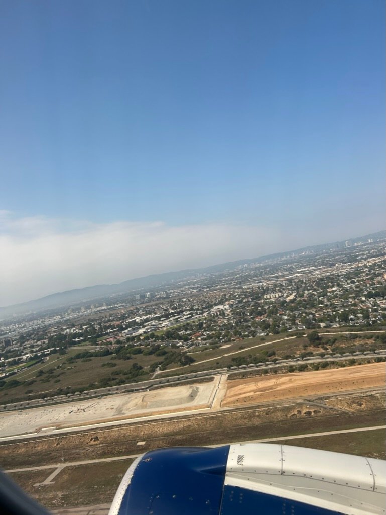 View from airplane window during landing, showing a cityscape, construction zones, and a blue engine in the foreground.
