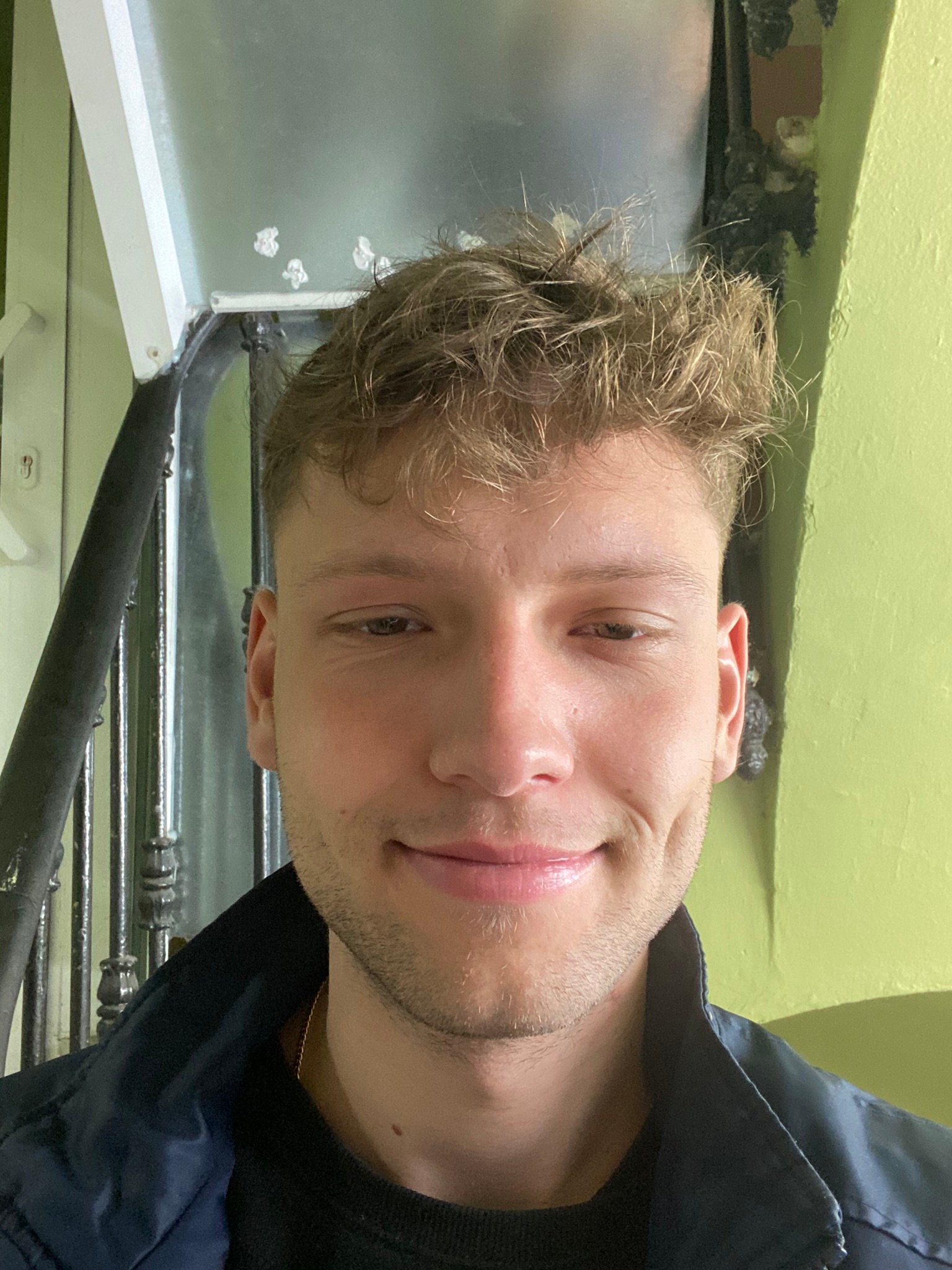 A young man with tousled light brown hair smiles softly while standing indoors in front of a green wall and a black spiral staircase