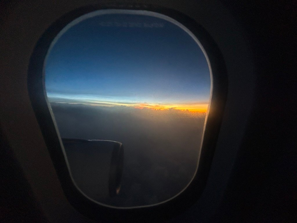 A view from an airplane window showing a sunset horizon above the clouds with a dark engine partially visible