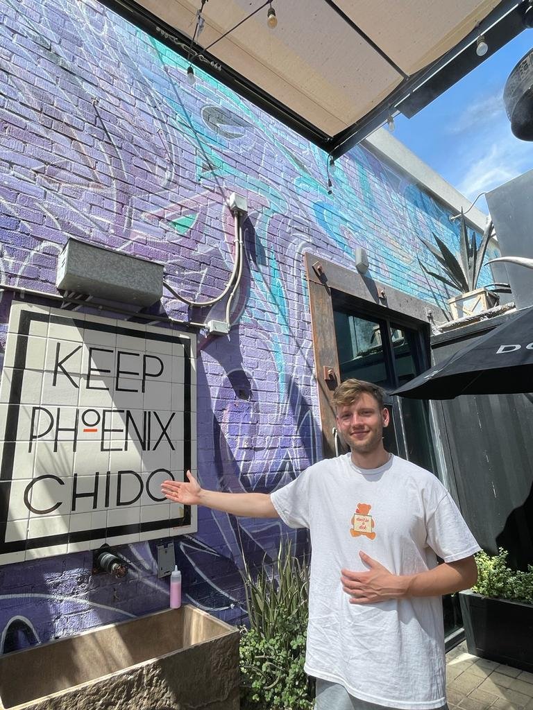 A smiling man in a white t-shirt points to a tiled sign reading "Keep Phoenix Chido" on a purple mural-covered wall in bright sunlight.