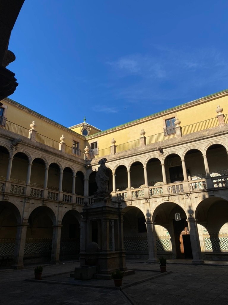 Stone courtyard with a central statue surrounded by arched hallways and balconies under a clear blue sky.