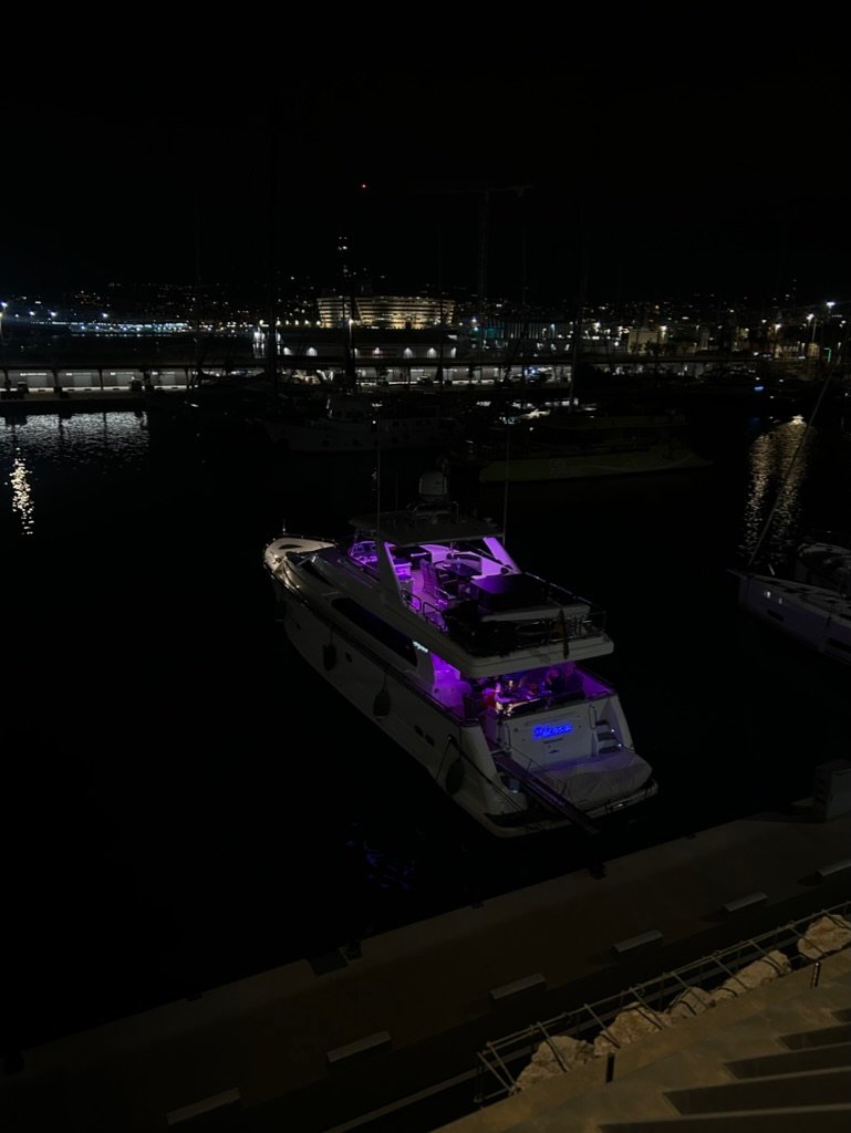 A luxury yacht with glowing purple lights docked at a marina at night, surrounded by dark water and distant city lights.