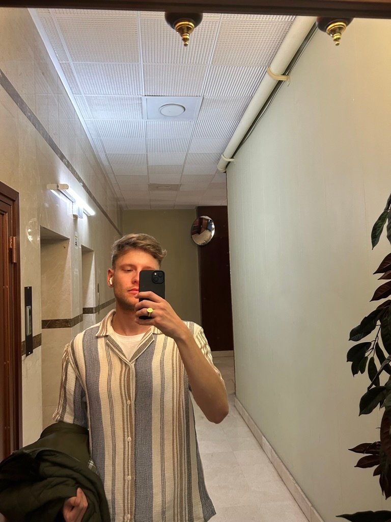 A man takes a mirror selfie in a hallway with tiled walls and ceiling, wearing a striped shirt and holding a jacket.