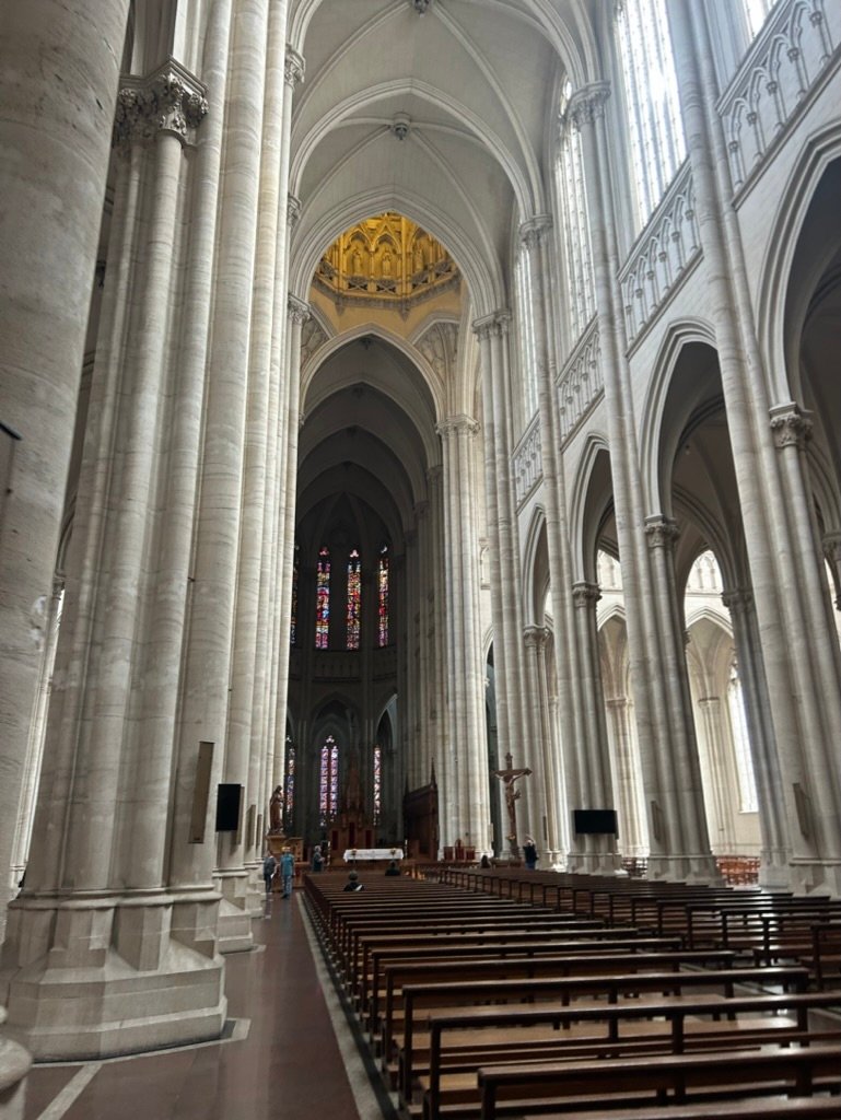The interior of a grand cathedral with towering white stone columns, arched ceilings, stained glass windows, and rows of wooden pews