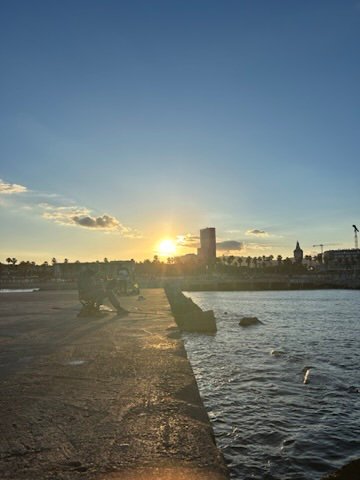 Two people sit at the edge of a concrete pier during sunset, with the sun casting golden light over the water and skyline.