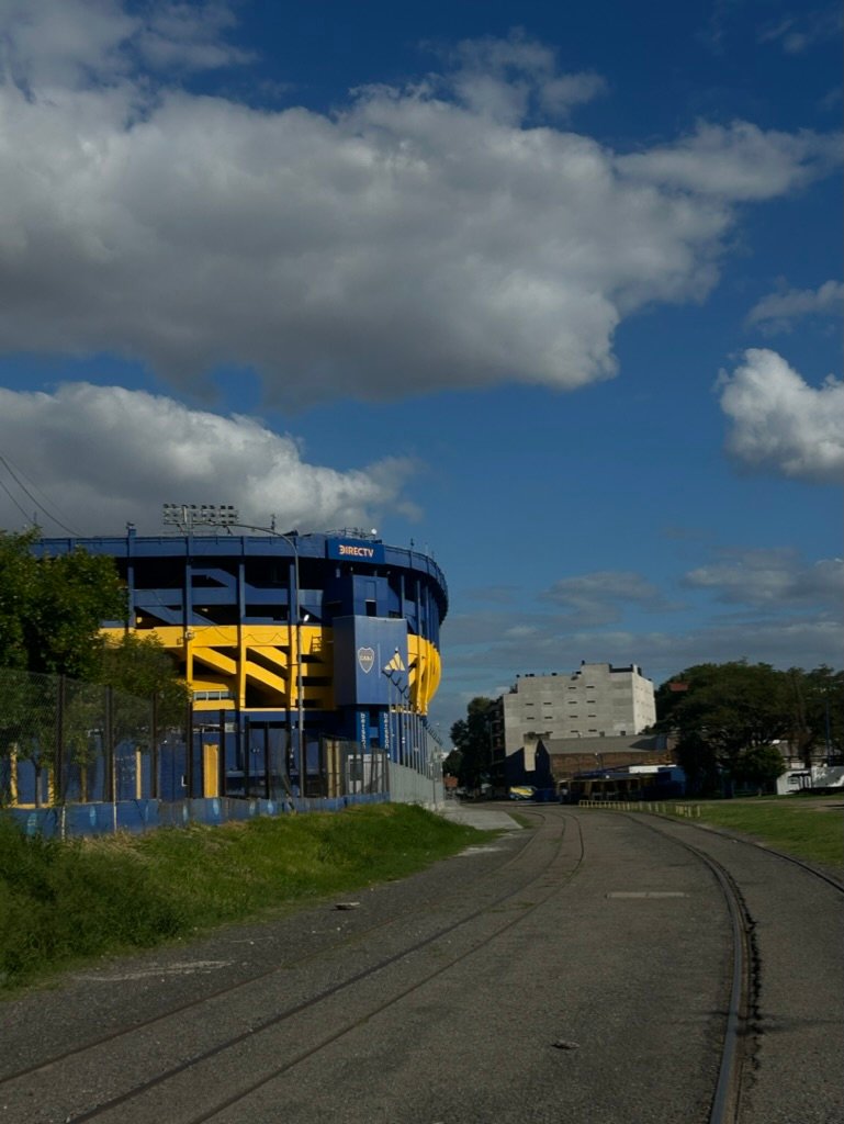 Blue and yellow stadium with a bold sky and railroad tracks curving toward it under fluffy clouds.