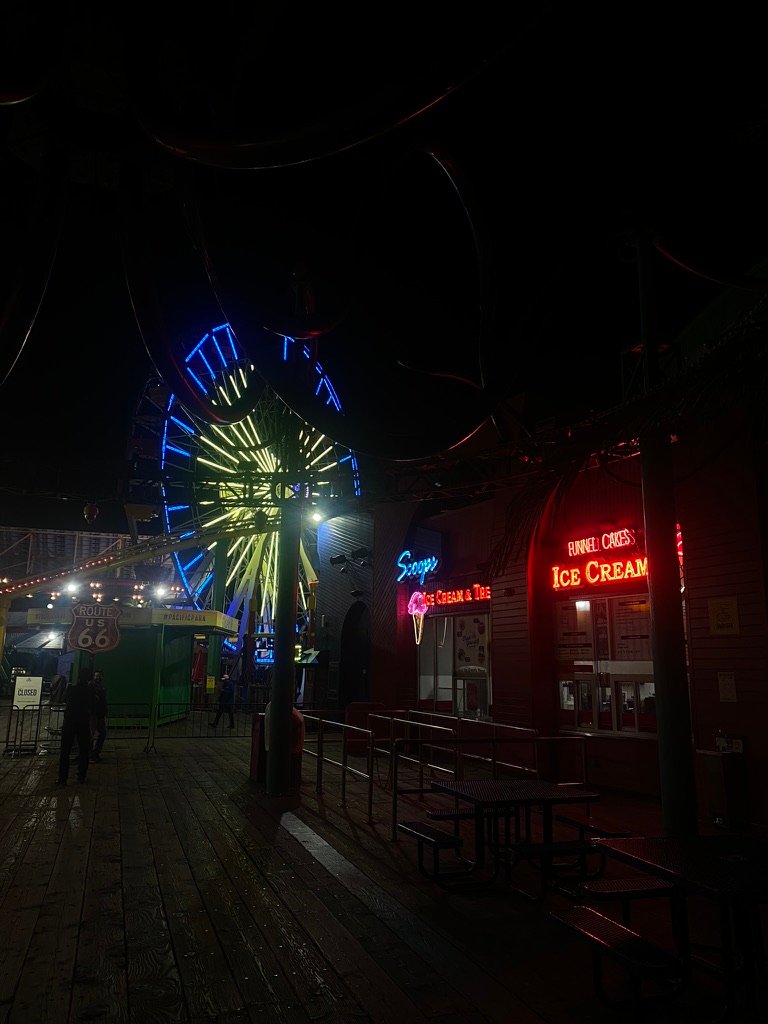 A nighttime view of a glowing Ferris wheel and neon-lit ice cream shop on a wooden boardwalk at Pacific Park.